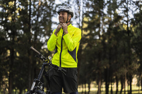 Mature man standing with bicycle and adjusting helmet in forest - JCCMF10896