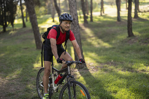 Mature man wearing helmet and riding bicycle in forest on sunny day - JCCMF10898