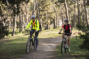 Mature friends wearing helmet and riding bicycles in forest on sunny day - JCCMF10900