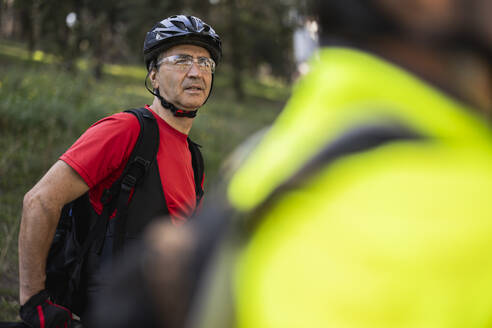 Mature man wearing cycling helmet and standing with friend in forest - JCCMF10901