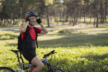 Smiling man sitting on bicycle and adjusting eyewear in forest - JCCMF10907