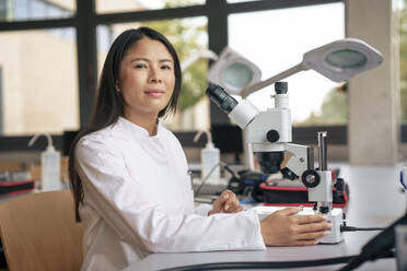 Lächelnde Wissenschaftlerin mit Mikroskop am Schreibtisch sitzend - JOSEF21867