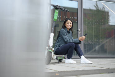 Glückliche Geschäftsfrau, die auf einem Elektroroller mit Smartphone auf dem Gehweg sitzt - JOSEF21954