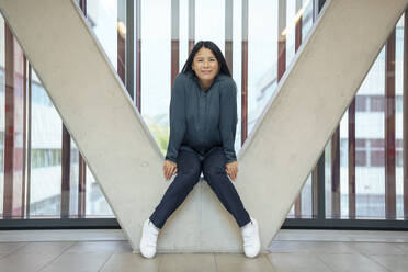 Lächelnde Geschäftsfrau, die auf einer architektonischen Säule im Büro sitzt - JOSEF21977