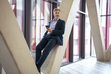 Thoughtful businessman with smart phone leaning on column - JOSEF21992