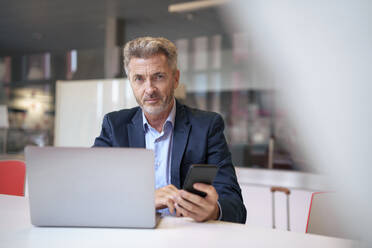 Smiling mature businessman sitting with mobile phone and laptop at table - JOSEF22011