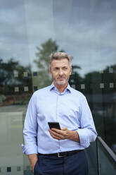 Businessman standing with mobile phone in front of glass - JOSEF22015