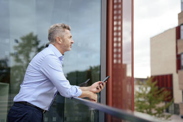 Thoughtful businessman leaning on railing with mobile phone in front of glass - JOSEF22016