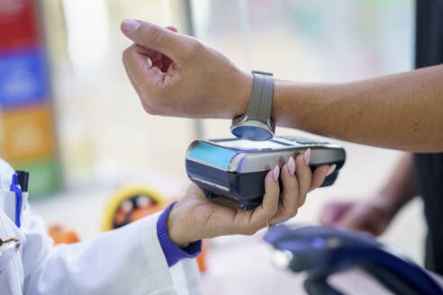 Hand eines Kunden, der in einer Apotheke per Tap-to-Pay-Verfahren über eine Smartwatch bezahlt - JSMF02922