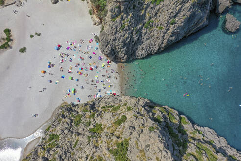 Luftaufnahme des Strandes Sa Calobra mit Menschen, die im klaren blauen Wasser schwimmen und mit Handtüchern und Sonnenschirmen auf dem felsigen Sand zwischen zwei Bergen auf den Balearen, Spanien, liegen. - AAEF23817