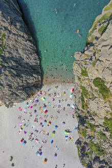 Luftaufnahme des Strandes Sa Calobra mit Menschen, die im klaren blauen Wasser schwimmen und mit Handtüchern und Sonnenschirmen auf dem felsigen Sand zwischen zwei Bergen auf den Balearen, Spanien, liegen. - AAEF23818