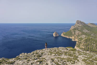 Luftaufnahme einer Frau, die auf einer hohen Klippe steht und auf den weiten Ozean mit Bergen im Hintergrund auf den Balearen, Spanien, blickt. - AAEF23820