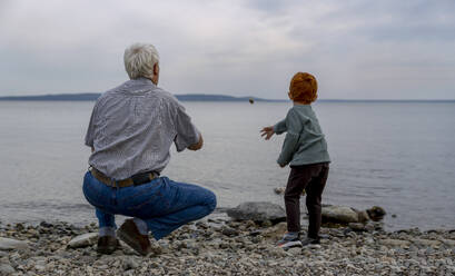 Großvater mit Enkel, der am Strand einen Kieselstein ins Meer wirft - MBLF00113