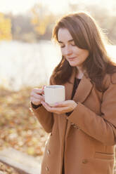 Glückliche Frau im Mantel und mit einer Tasse Tee in einem herbstlichen Park - ONAF00678