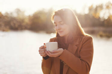 Glückliche Frau mit einer Tasse Tee in der Nähe eines Sees - ONAF00679