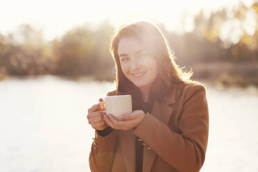 Lächelnde Frau hält eine Tasse Tee in der Nähe eines Sees bei Sonnenuntergang - ONAF00680