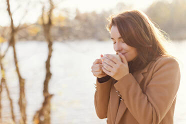 Smiling woman drinking tea from cup by lake at sunset - ONAF00681