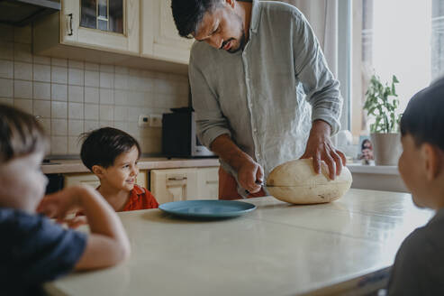 Vater schneidet Melone mit Kindern am Tisch in der Küche - ANAF02396