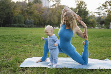 Blond mother doing stretching exercise with toddler son in park - YBF00285