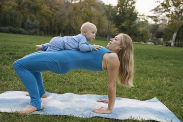 Blond mother balancing son and doing yoga in park - YBF00291