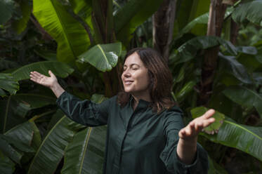 Smiling woman standing with arms raised in botanical garden - VRAF00220