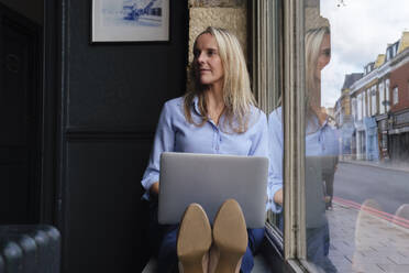 Blonde Freiberuflerin sitzt mit Laptop auf der Fensterbank in einem Cafe - ASGF04711