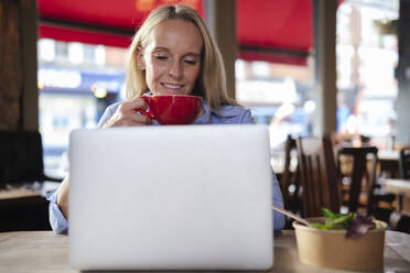 Lächelnder Freiberufler, der eine Kaffeetasse hält und an einem Laptop in einem Café arbeitet - ASGF04712