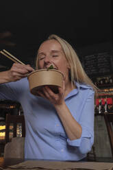 Blond woman eating food with chopsticks at table in pub - ASGF04714