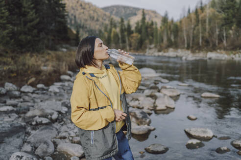 Woman drinking water from bottle by river in forest - VBUF00505