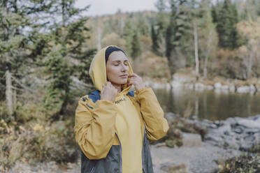 Woman wearing hooded shirt and standing by river in forest - VBUF00508