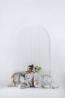 Junger Mann und Frau mit Virtual-Reality-Headset sitzen Rücken an Rücken vor einer weißen Wand - YTF01350