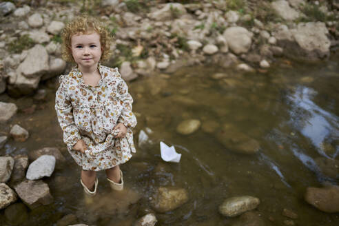 Lächelnde niedliche Mädchen stehen in der Nähe von Papier Boot und Felsen in Wasser Pfütze - ANNF00588