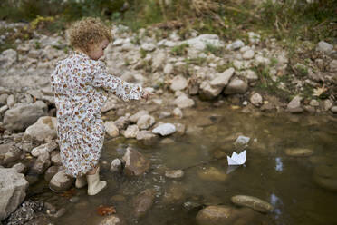 Cute girl playing and pointing on paper boat in water puddle near rocks - ANNF00589