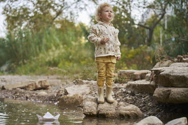 Smiling girl standing on rock with paper boat in water puddle - ANNF00598