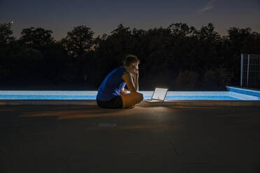 Freiberufler arbeitet nachts am Laptop in der Nähe des Schwimmbads - MAMF02900