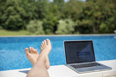 Foot of woman near laptop at poolside on sunny day - MAMF02904