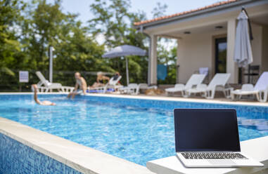 Laptop mit leerem Bildschirm in der Nähe des Schwimmbads an einem sonnigen Tag - MAMF02910