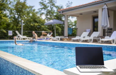 Laptop with blank screen kept near swimming pool on sunny day - MAMF02910