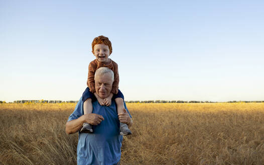 Glücklicher Großvater trägt Enkel auf den Schultern in einem Weizenfeld unter dem Himmel - MBLF00137