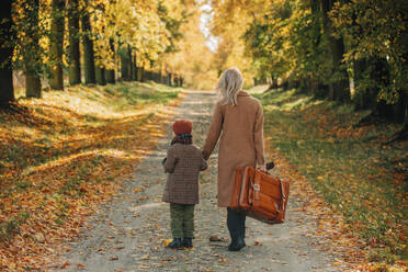 Boy walking with mother holding suitcase at autumn park - VSNF01417