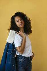 Woman holding denim jacket and standing against yellow background - EBSF04024