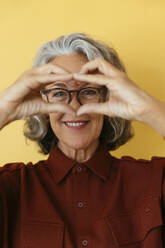 Lächelnde ältere Frau zeigt Herzzeichen vor gelbem Hintergrund - EBSF04029