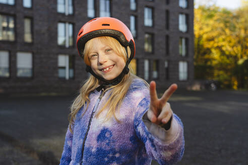 Smiling girl wearing helmet gesturing peace sign with hand - IHF01832