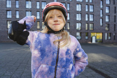 Lächelndes Mädchen mit Schutzausrüstung, das vor einem Gebäude seine Muskeln spielen lässt - IHF01835