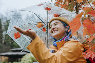 Smiling woman holding umbrella and enjoying rain in autumn park - YTF01377