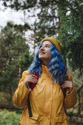 Lächelnde Frau, die einen Rucksack trägt und in der Nähe eines Baumes im Wald steht - YTF01408