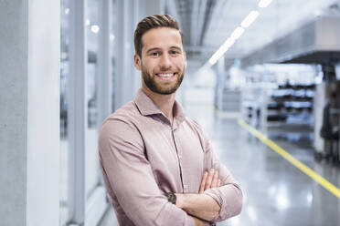 Smiling young businessman with arms crossed standing in factory - DIGF20984