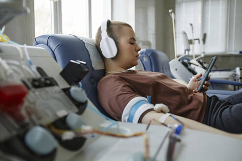 Young woman listening to music through headphones and donating blood - KPEF00312
