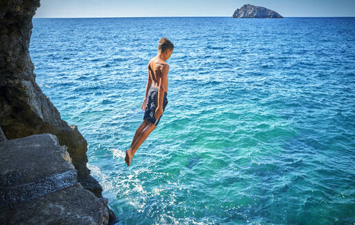 Boy jumping from cliff in blue sea - DIKF00771