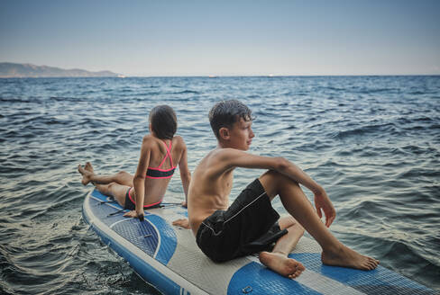 Brother and sister sitting on paddle board in sea - DIKF00775